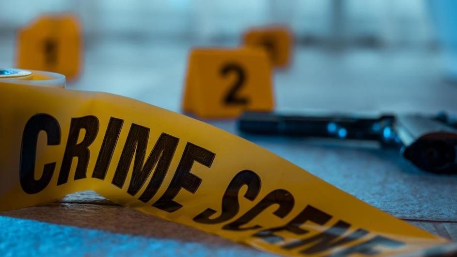 Image of crime scene tape in the foreground and a gun lying on its side in the background.
