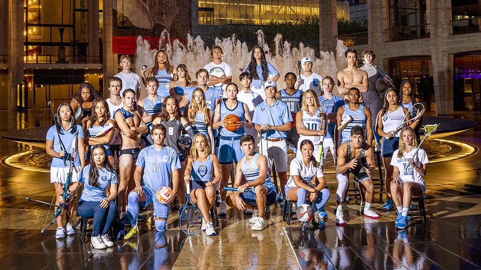 33 Columbia University student-athletes in light blue team uniforms pose in front of Revson Fountain at Lincoln Center for the Performing Arts at night in New York City. 