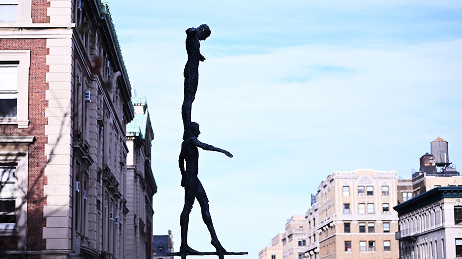 The bronze Tightrope Walker statue by Kees Verkade of one walker on the shoulders of another on Revson Plaza against a blue sky at Columbia University. 