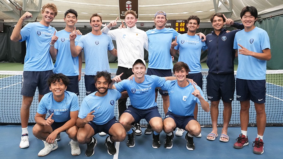 Twelve men's tennis players in Columbia blue uniforms smile and pose with their index fingers up to show that they are Ivy League champions.