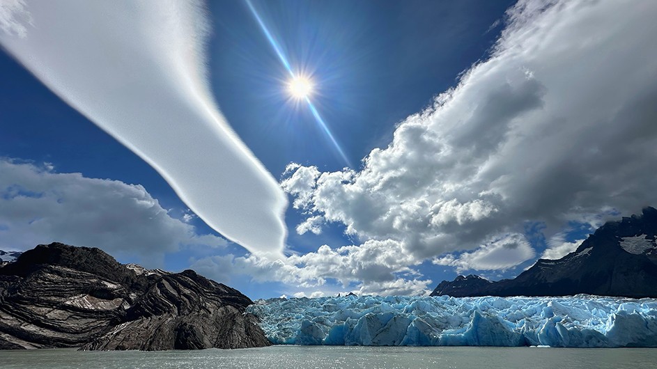 Long cloud formations and bright sun in blue sky above icy glacier and lake at Grey Glacier and Grey Lake in Torres del Paine National Park in Chile.