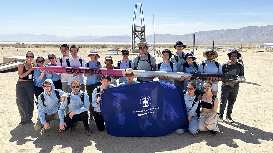 Several Columbia University students in light blue shirts pose while holding a long hybrid rocket on a sunny day in the Mojave Desert. 