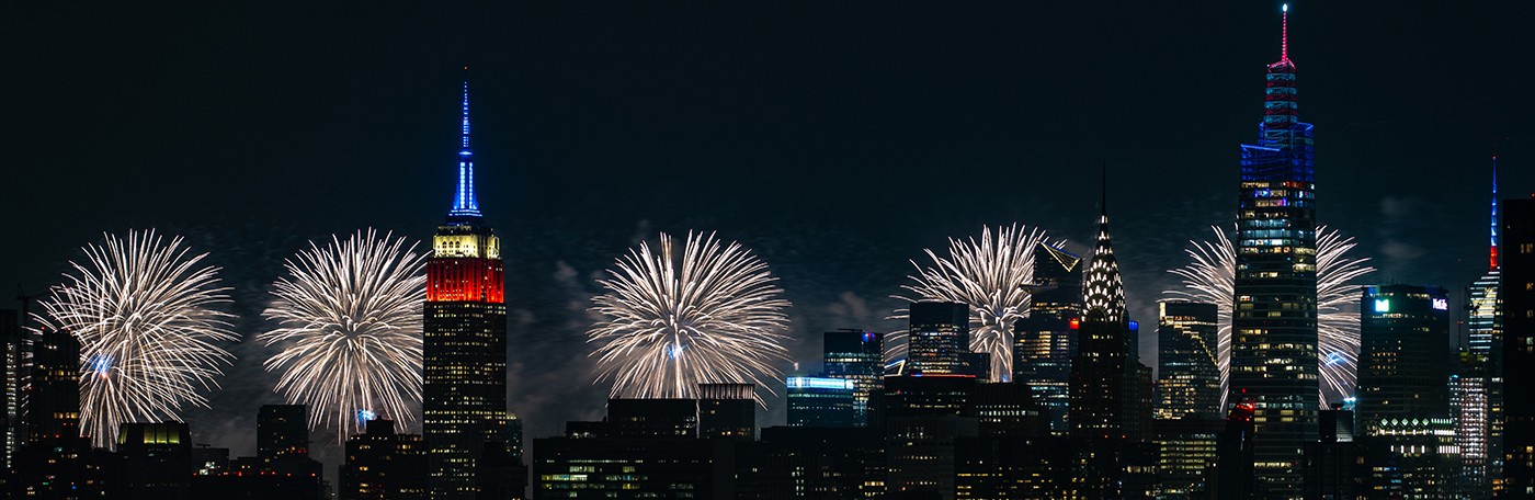 New York City skyline of Empire State building, Chrysler Building, and other skyscrapers with white fireworks illuminating the sky on July 4th. 