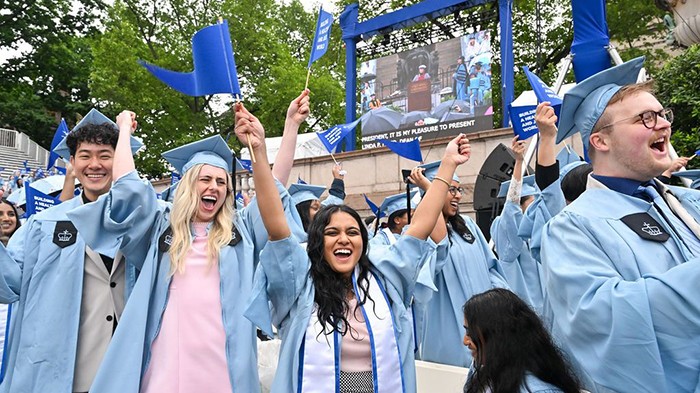 "Building a healthy and just world," read the flags of Mailman School of Public Health graduates. Photo by Diane Bondareff. Mailman School of Public Health graduates celebrate with blue flags waving in the air.
