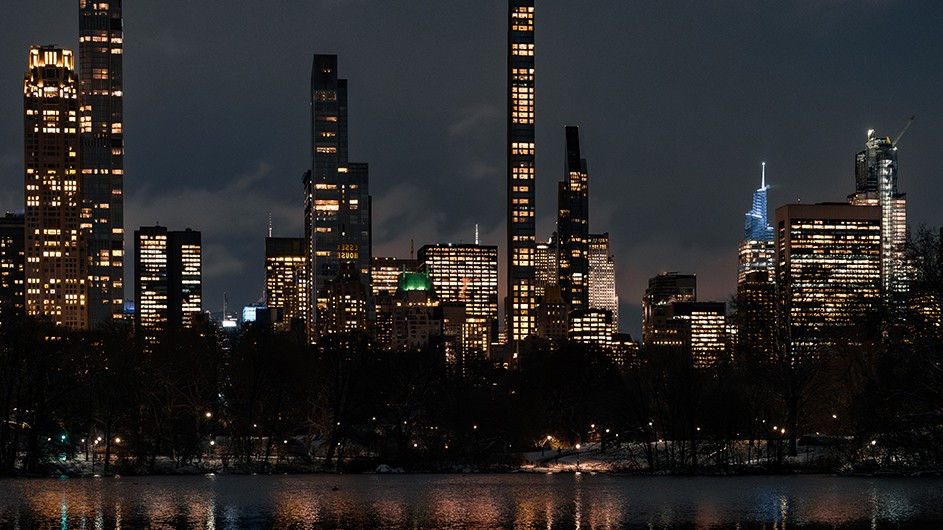 Illuminated midtown Manhattan skyscrapers reflected on Central Park's pond lined by snowy tree-lined paths on a winter night. 