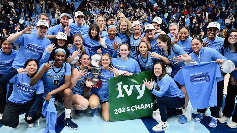 Several university students and coaches in light blue Columbia basketball uniforms smile as they hold up three fingers and hold a trophy and signage for Ivy 2025 Champions in front of fans in Levien Gym at Columbia University.  