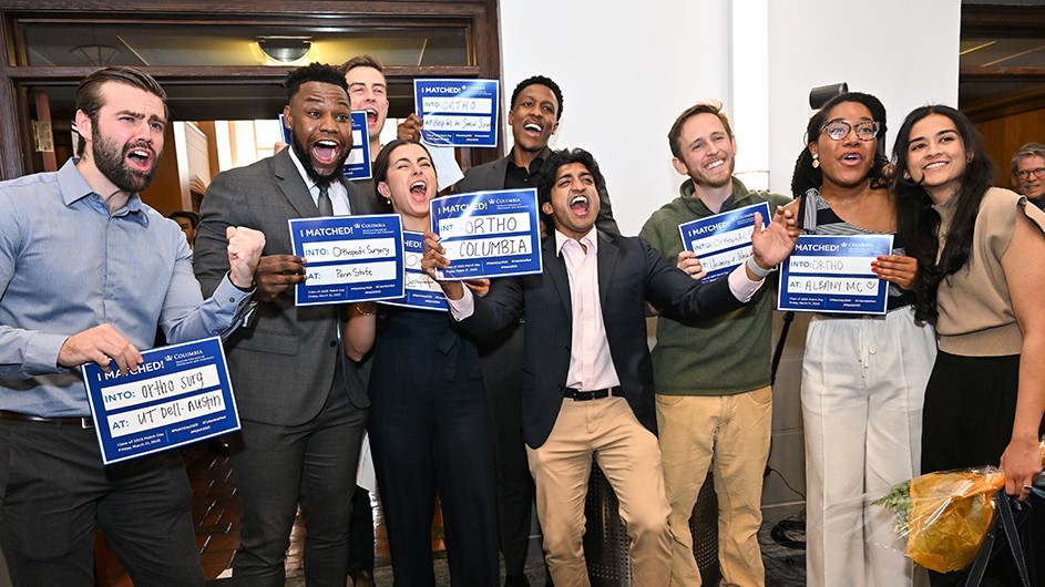 Nine university students celebrate and pose with blue signs during Columbia's Vagelos College of Physicians & Surgeons' annual Match Day for residencies at Columbia Medical Center.