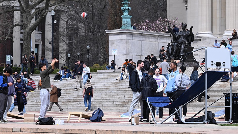 University students gathered around Alma Mater statue at Columbia University, some engaging in activities like taking photos and talking, with a few sitting on the steps. A man in the foreground shoots a basketball into a hoop.