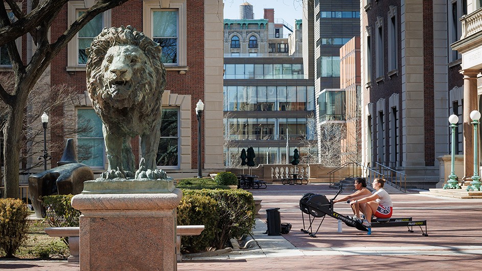 Two university students row on stationary rowing machines near Scholars’ Lion bronze sculpture in front of Havemeyer Hall and Math Hall at Columbia University on a sunny day. 