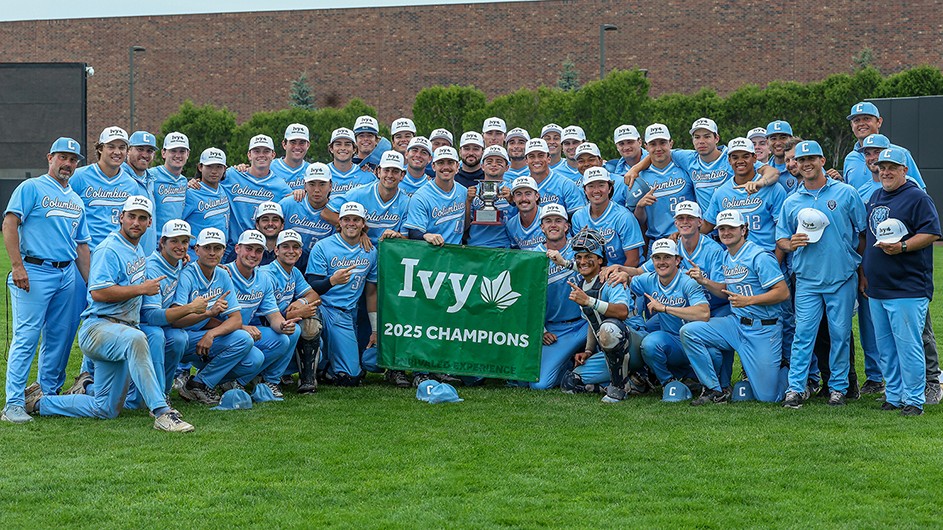 Several men's baseball players and coaches in Columbia blue uniforms smile and pose with a trophy and Ivy League 2025 Champions sign at Columbia's Baker Athletics Complex.