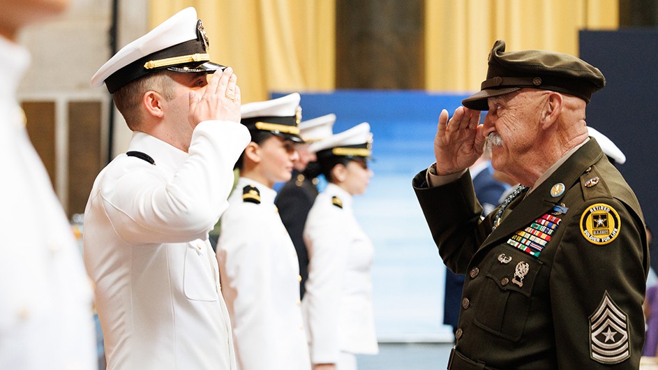 A university student in white naval uniform salutes a retired army veteran, who returns a salute, alongside other university students in uniform at Columbia University's Naval ROTC commissioning ceremony. 