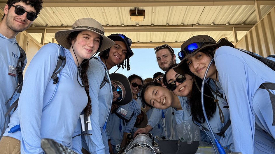 Several Columbia University students in light blue shirts smile and pose with their hybrid rocket in a shed in the Mojave Desert. 