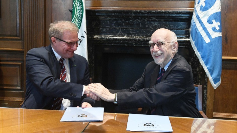 Two elderly white men holding hands and smiling in front of flags