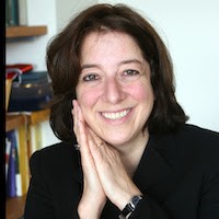 Anya Schiffrin, a woman with brown hair, in a black blazer with her hands clasped by her face, smiling.