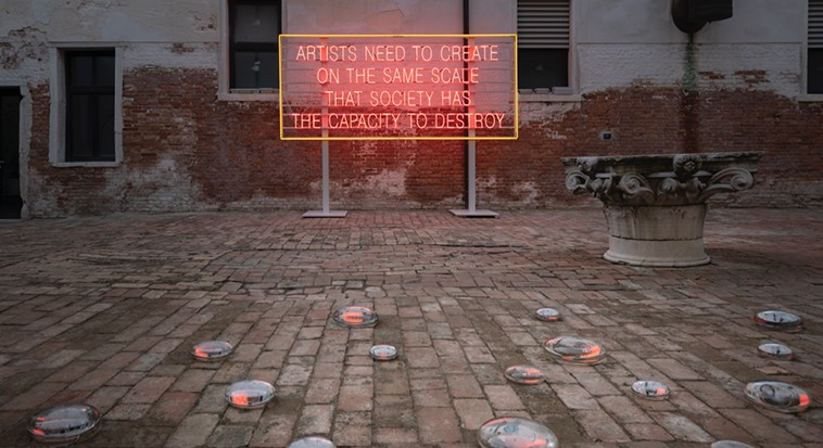 Lauren Bon and the Metabolic Studio, Artists need to Create on the Same Scale that Society Has the Capacity to Destroy, 2019 Courtesy the artist. Neon sign states: Artists need to create on the same scale that society has the capacity to destroy. Below are a series of glass disks placed on a brick pavement.
