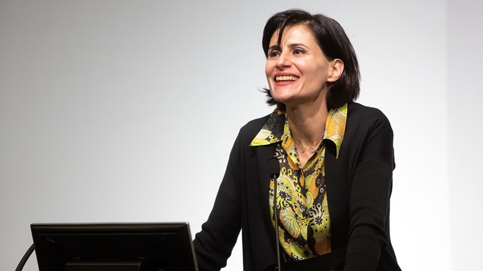 Amale Andraos, Dean of the School of Architecture, Planning and Preservation. A woman in a yellow flowered shirt and black sweater standing in front of a computer at a lectern.