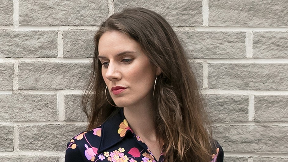 Leslie Jamison: a woman with long brown hair in a flowered shirt standing in front of a gray brick wall.
