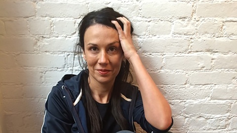 A woman in jeans sitting on a wood floor in front of a white brick wall.