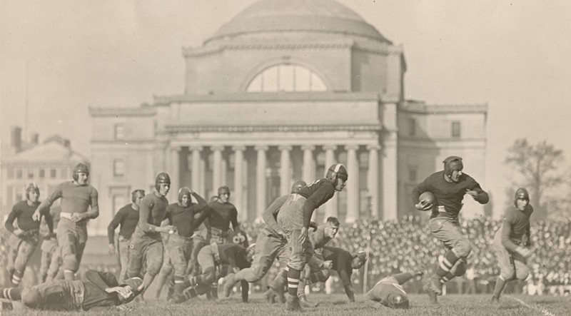 1921 football game on South Field, Columbia vs. NYU. A group of men wearing old-fashioned uniforms and playing football in front of a classical building with steps.