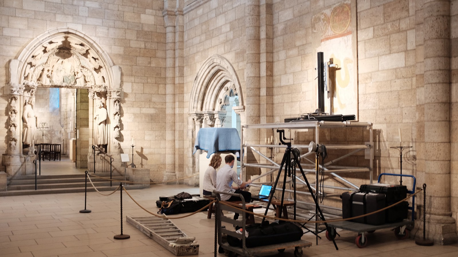 Two people working with equipment in a brick-walled medieval cloister.