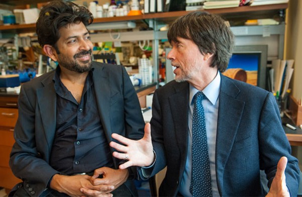 Prof. Siddhartha Mukherjee dressed in dark blazer and shirt sits with hands folded while speaking with Ken Burns dressed in dark blazier light blue shirt with blue tie in the reaer are shelves of books and picture frames.