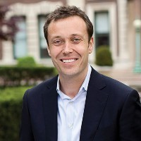 A man with closely cropped brown hair, smiling, and wearing a white buttoned-down shirt and dark suit jacket