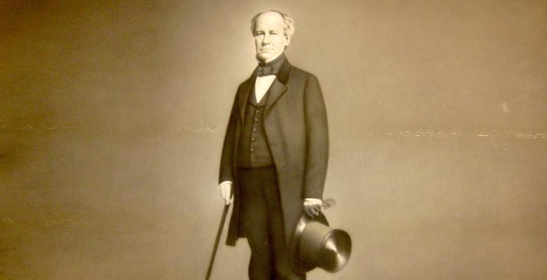 A black and white vintage image of Charles Anthon stands tall dressed in a dark suit with a long blazer jacket while holidng a top hat and walking cane.