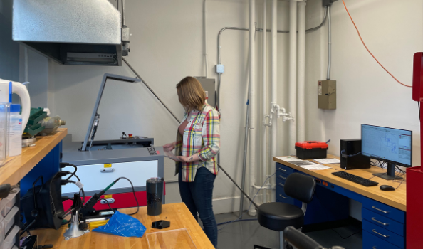 A laser cutter is another modern option to build computer-aided designs. Clara Wilson standing in front of laser cutter, with a computer behind her.