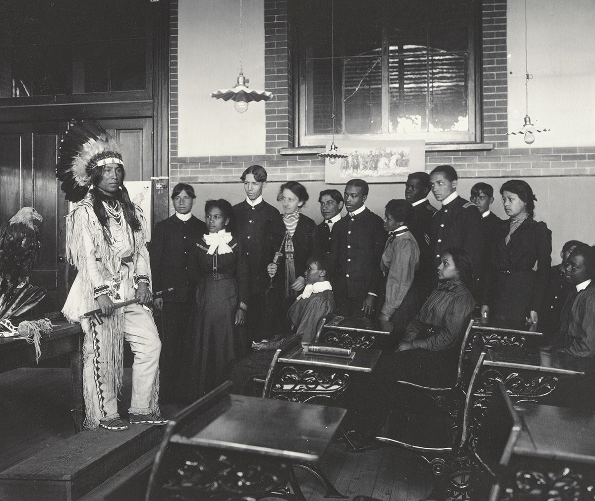 "Class in American History," the Hampton Album; platinum print by Frances B. Johnson. A Native American man wearing a feathered headdress stands in a classroom of students.