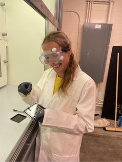 Doroshevich carefully cleans a newly fabricated resonator chip. Lana Doroshevich in a white lab coat, goggles, and black glove preparing a superconducting resonator chip.