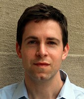 david pozen wearing a light blue colored collared button down shirt standing against a teal colored wall.  His hair is black