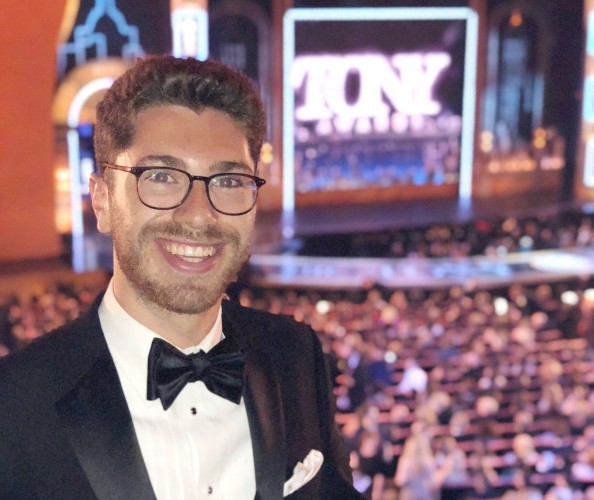 Treatman at the 2018 Tony Awards. David Treatman: A man with dark hair and glasses, wearing a tux is smiling with a stage and crowds in the background.