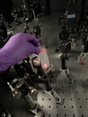 A purple gloved hand holds a card in front of a red laser beam on an lab table. 