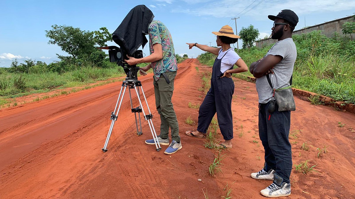 School of the Arts film student Olive Nwosu on set in Nigeria, shooting her short movie 'Troublemaker.' Three people standing on a red-dirt road, surrounded by grass and blue sky.