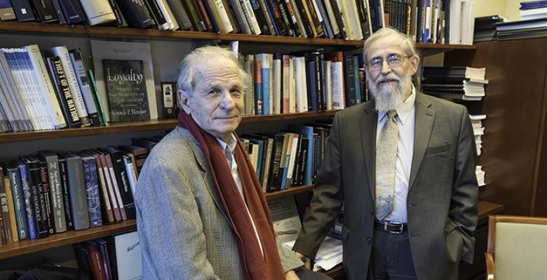 George Fletcher wearing tan blazer with a brown colored scarf draped around his neck with a white shirt stands with Saul Berman who is dressed in a brown colored suite with a tan necktie and white shirt both men are standing in front of a wall of shelved books