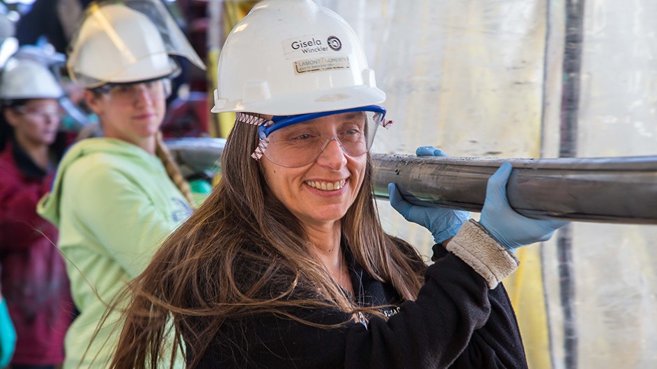 Gisela Winckler and crew carry sediment cores on the catwalk of drill ship Joides. Photo by Tim Fulton, IODP Smiling woman leading team in white hard hats on drill ship carrying think grey tubing