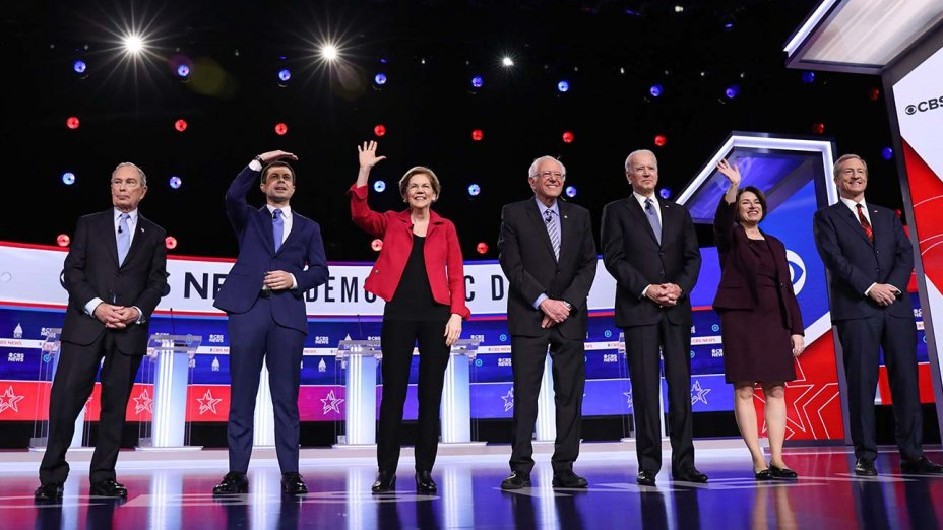 Seven people trying for the Democratic presidential nominations standing on red, white and blue decorated stage. Some of them are waving to the audience.