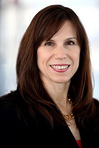 Jennifer Rodgers dressed in a dark blazer smiles directly into camera lens. Hair is dark a dark auburn shoulder length bob cut with parted bangs.