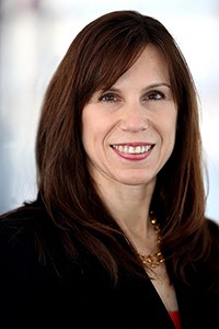 Jennifer Rodgers dressed in a dark blazer smiles directly into camera lens. Hair is dark a dark auburn shoulder length bob cut with parted bangs. 