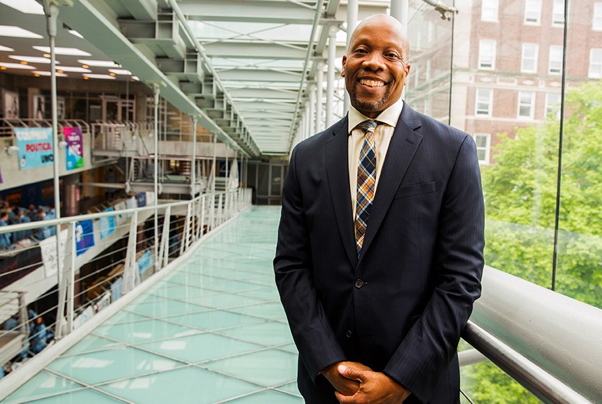 Lance Freeman Lance Freeman in a dark gray suit smiling, standing on a glass ramp that looks over a tree on the right and other floors on the left.