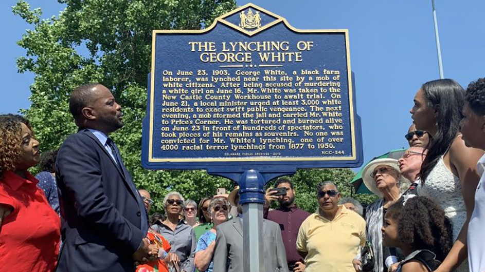 Sen. Darius Brown, Savannah Shepherd and others read the Lynching of George White Historical Marker just minutes after it was unveiled at Greenbank Park, DE, on June 23, 2019. Shepherd spearheaded the effort to have the marker erected. (Credit: Scott Goss/Delaware Senate Majority Caucus) A group of people including African Americans gathered around a plaque