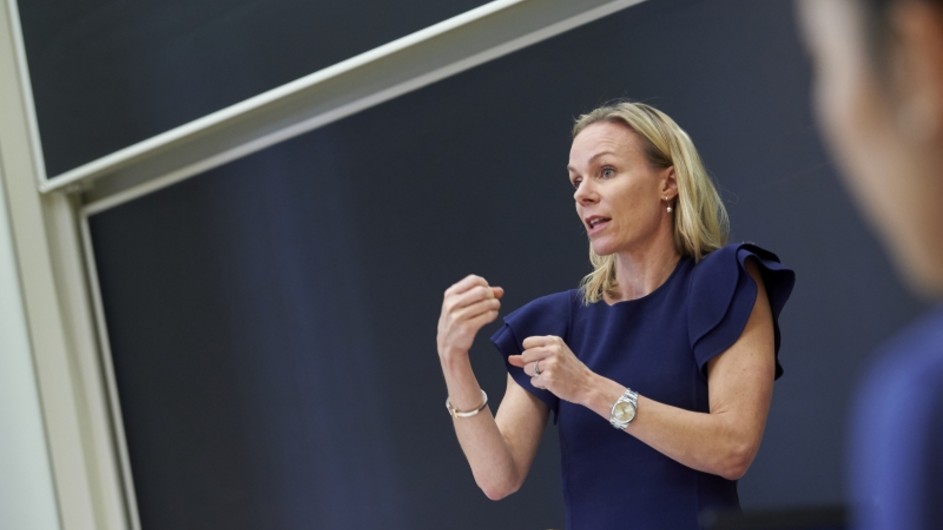 A blond woman speaking in front of a black board