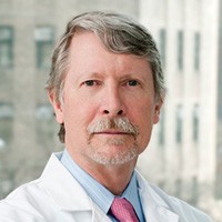 A grey-haired man with mustache and beard, wearing blue button-down shirt, pink tie and white lab coat.
