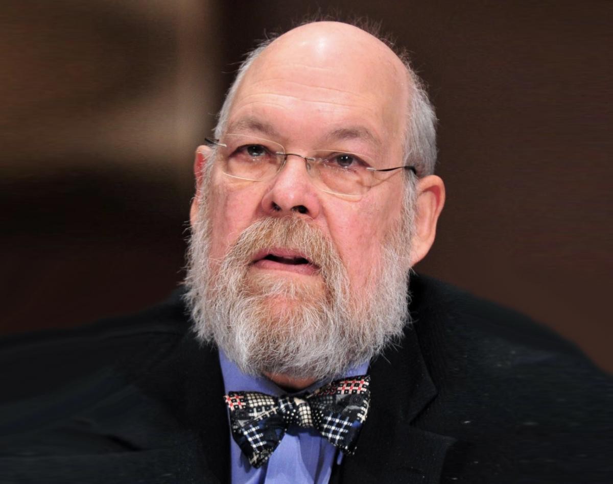 Michael Graetz dressed in a dark blazer blue collared shirt with a blue red, white and blue bowtie. Wear clear rimmed eyeglasses salt and pepper beard looking straight into camera.