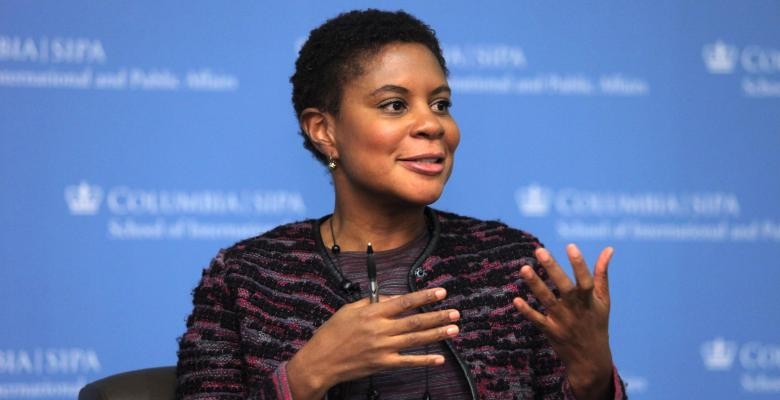 A woman with close-cropped hair smiles and gestures as she speaks to someone off-camera