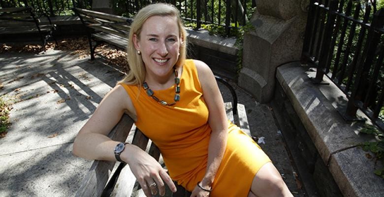 A woman sites on a park bench in an orange sleeveless dress, smiling into the camera.