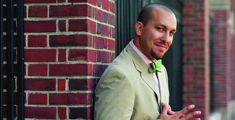 Photo by Gabriel Cooney A man leans against a brick column, wearing a suit and bow tie, smiling into the camera