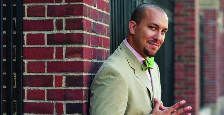 A man leans against a brick column, wearing a suit and bow tie, smiling into the camera