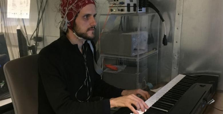 A man plays a keyboard wearing on his head a tangle of wires connected to a bank of computer equipment.