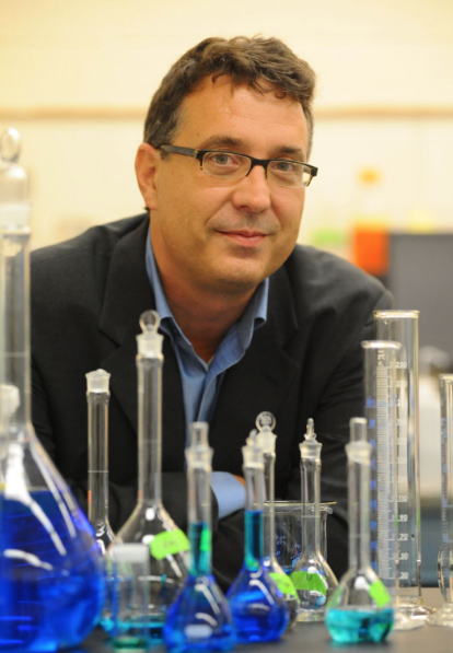 Alan West. Photo by Eileen Barroso A man, smiling and wearing glasses, sits a a table filled with beakers holding brightly colored liquids